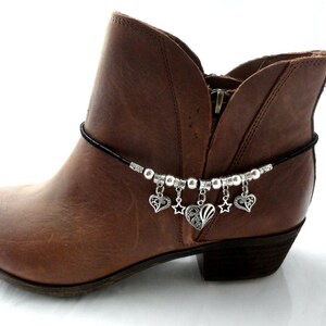 May include: A brown leather ankle boot with a black cord ankle bracelet. The bracelet features silver-colored beads and charms, including hearts and stars. The boot has a zipper and a low heel.