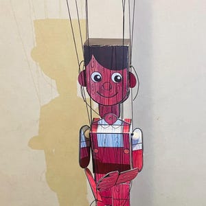 Könnte beinhalten: Handgefertigte Holzmarionette in Rot- und Brauntönen. Die Puppe hat ein lächelndes Gesicht, dunkles Haar und trägt eine Latzhose. Sie hält ein rotes Objekt und ist an schwarzen Fäden aufgehängt.