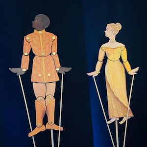 Pode incluir: Dois fantoches de papel em varinhas. O fantoche à esquerda é um homem de pele morena vestindo uma roupa laranja e dourada. O fantoche à direita é uma mulher de cabelo loiro vestindo um vestido amarelo.