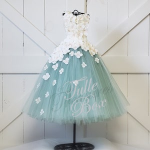 May include: A sage green tulle dress with white flower embellishments. The dress is on a mannequin and has a full skirt.