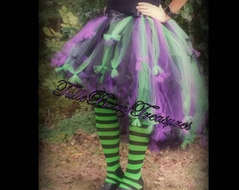 May include: A purple and green tulle tutu skirt with black ribbon bows. The skirt is worn with green and black striped tights and black shoes.