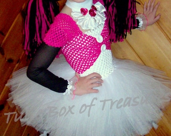 May include: A young girl dressed as a doll for Halloween. She is wearing a pink and black yarn wig, a pink and white crocheted top, a white tulle tutu, and black tights. The tutu has white tassels hanging from the bottom. She is also wearing black shoes and has a heart-shaped sticker on her cheek.