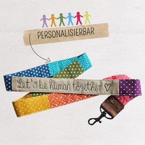 Könnte beinhalten: Ein farbenfrohes Stoffband mit dem gestickten Text "Let's be human together". Das Band hat ein braunes Lederband mit einem Metallclip. Der Text "PERSONALISIERBAR" ist im Hintergrund sichtbar.