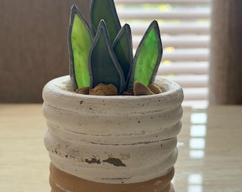 May include: A handcrafted, decorative succulent plant in a small, white and tan ceramic pot. The plant features green glass leaves with dark outlines, and small brown stones at the base. The pot has a textured, ribbed design.