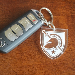 Puede incluir: Una llave de coche negra con un logotipo de Mazda plateado y tres botones. La llave está unida a un llavero de acrílico transparente con un logotipo blanco de casco y espada espartano.