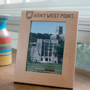 May include: A wooden picture frame with a photo of a building and the text "ARMY WEST POINT" engraved on the top. The frame is light brown and has a simple design.