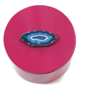 May include: A polished, round, magenta-colored container with a natural blue and white agate slice on top. The agate has concentric bands of color and a natural, organic shape. The container is set against a white background.