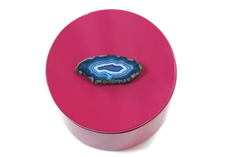 May include: A polished, round, magenta-colored container with a natural blue and white agate slice on top. The agate has concentric bands of color and a natural, organic shape. The container is set against a white background.