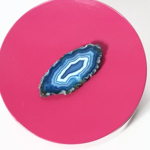 May include: A polished, oval-shaped agate stone with concentric bands of blue and white, resting on a bright pink circular surface. The agate's natural patterns are clearly visible, showcasing its unique geological formation. The stone is a decorative item.