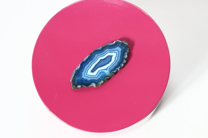 May include: A polished, oval-shaped agate stone with concentric bands of blue and white, resting on a bright pink circular surface. The agate's natural patterns are clearly visible, showcasing its unique geological formation. The stone is a decorative item.