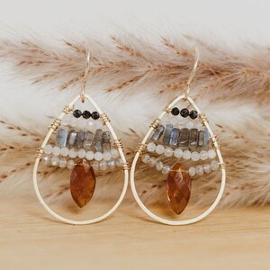 May include: Gold-toned teardrop earrings with intricate beadwork. The earrings feature a combination of white, gray, and black beads, along with a large, amber-colored gemstone at the bottom. The design is suspended from a gold hook.