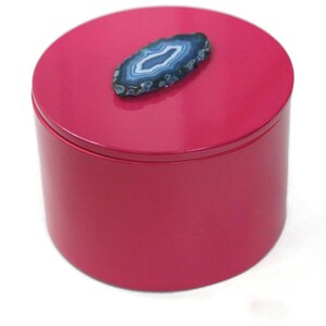 May include: A cylindrical, magenta-colored box with a matching lid. A blue and white agate stone is affixed to the top of the lid. The box is likely used for storing jewelry or small items.