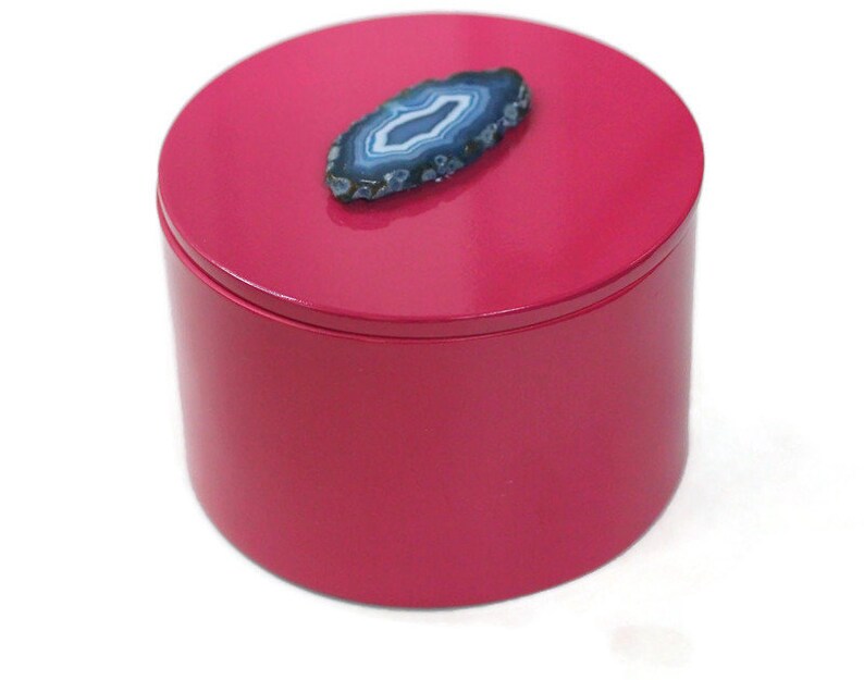 May include: A cylindrical, magenta-colored box with a matching lid. A blue and white agate stone is affixed to the top of the lid. The box is likely used for storing jewelry or small items.