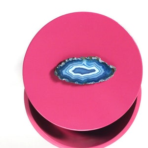 May include: A polished, oval-shaped agate stone with concentric bands of white, blue, and teal hues rests on a bright pink, circular surface. The stone's edges are rough and textured, contrasting with the smooth surface. The background is white.