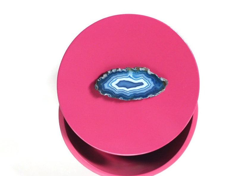 May include: A polished, oval-shaped agate stone with concentric bands of white, blue, and teal hues rests on a bright pink, circular surface. The stone's edges are rough and textured, contrasting with the smooth surface. The background is white.
