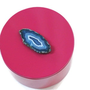 May include: A round, hot pink box with a polished finish. A blue and white agate slice is placed on top. The agate has a natural, layered pattern with shades of blue and white. The box is set against a white background.