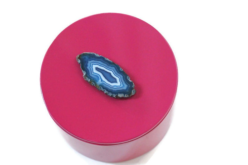 May include: A round, hot pink box with a polished finish. A blue and white agate slice is placed on top. The agate has a natural, layered pattern with shades of blue and white. The box is set against a white background.