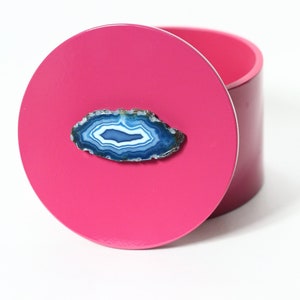 May include: A magenta-colored round box with a matching lid. The lid is adorned with a blue and white agate slice. The box is cylindrical and sits on a white surface.