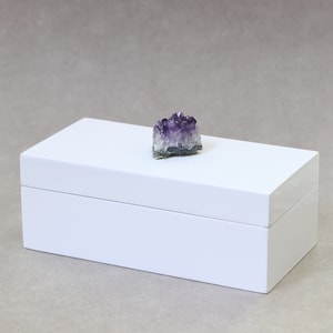 May include: A white rectangular box with a glossy finish, topped with a cluster of purple amethyst crystals. The box is closed and sits on a neutral-toned surface, creating a minimalist aesthetic. The amethyst adds a natural contrast to the clean white box.