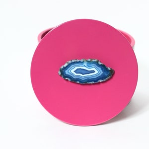 May include: A bright pink, round coaster with a polished, blue and white agate slice affixed to the surface. The agate has concentric bands of color, creating a natural, decorative element. The coaster is set against a white background.