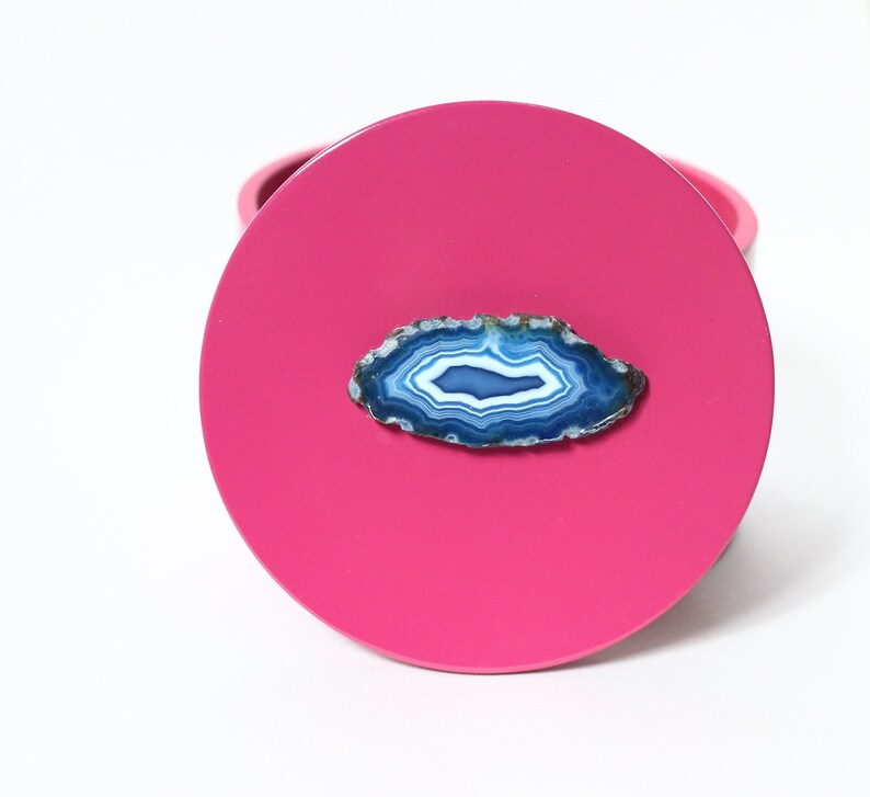 May include: A bright pink, round coaster with a polished, blue and white agate slice affixed to the surface. The agate has concentric bands of color, creating a natural, decorative element. The coaster is set against a white background.