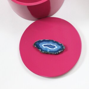 May include: A vibrant fuchsia pink lid with a polished finish, paired with a section of a matching container. A polished, oval-shaped blue agate stone with white and blue banding rests on the lid. The agate has a natural, textured edge.