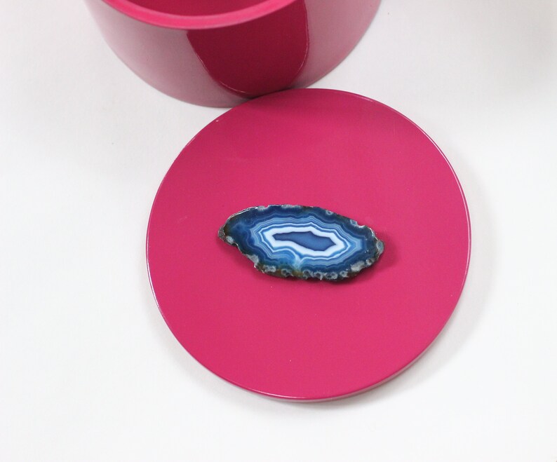 May include: A vibrant fuchsia pink lid with a polished finish, paired with a section of a matching container. A polished, oval-shaped blue agate stone with white and blue banding rests on the lid. The agate has a natural, textured edge.