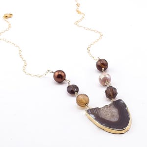 May include: A gold chain necklace featuring a pendant with a dark brown and gray agate slice with a gold-colored trim. The necklace also includes a series of brown, tan, and clear beads.