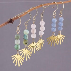 Peut inclure: Plusieurs boucles d'oreilles dorées avec des perles. Chaque boucle d'oreille est ornée d'un motif de soleil doré et de trois perles vertes, roses ou bleues. Les boucles d'oreilles sont présentées sur une fine branche brune sur fond gris.