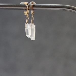 May include: A pair of gold-colored hoop earrings with clear, crystal-like pendants. The crystals are rectangular with irregular edges, suspended from the hoops by delicate gold wire. The background is a solid, muted gray.