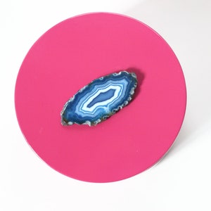 May include: A vibrant, overhead shot of a hot pink, circular object with a polished surface. A blue and white agate slice is centered on the pink surface. The agate has a natural, layered pattern.
