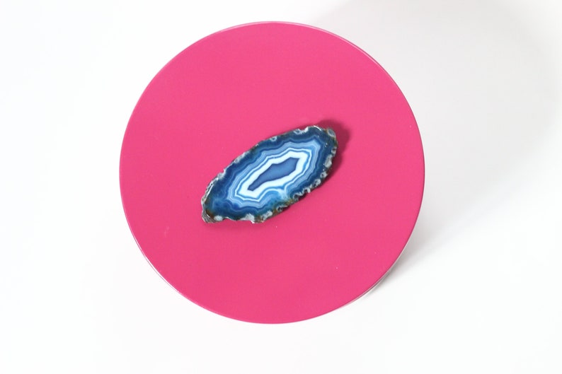 May include: A vibrant, overhead shot of a hot pink, circular object with a polished surface. A blue and white agate slice is centered on the pink surface. The agate has a natural, layered pattern.