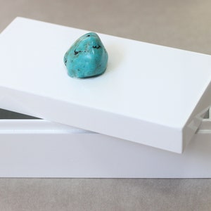 May include: A white rectangular box with a slightly ajar lid, showcasing a vibrant turquoise stone. The box has a glossy finish, and the stone is a natural, irregular shape. The overall aesthetic is minimalist and elegant.
