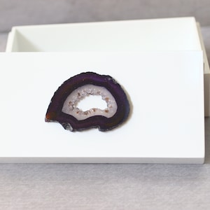 May include: A white rectangular box with an open lid, displaying a polished, sliced geode. The geode features a dark purple outer band, a white and light brown center, and a crystalline interior.