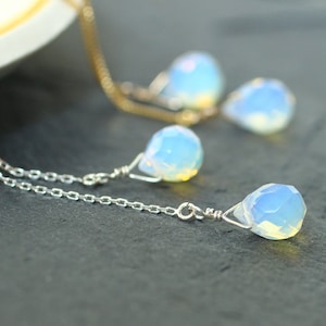 May include: A silver necklace with a delicate chain and a teardrop-shaped, opalescent pendant. The pendant has a faceted surface that catches the light, creating a shimmering effect. The necklace is displayed on a dark surface.