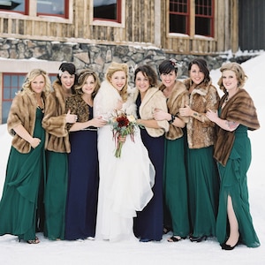 Pode incluir: Um grupo de damas de honra em vestidos verde esmeralda e capas de pele estão com a noiva em um vestido branco e uma capa de pele. Todas estão sorrindo e olhando para a câmera. As damas de honra estão usando diferentes estilos de capas de pele, incluindo estampa de leopardo e marrom sólido.