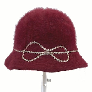 May include: A burgundy cloche hat with a soft, fuzzy texture. A sparkling rhinestone bow detail adorns the front. The hat is displayed on a clear, adjustable stand, showcasing its shape and design.