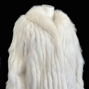 Peut inclure: Un manteau de fourrure blanc avec un large col et des manches longues, présenté sur un mannequin. Le manteau est mis en valeur sur un fond noir. Le texte "find your dream fur at MOXIEFURS.COM" est visible en bas.