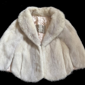 May include: A cream-coloured fur capelet featuring a wide collar and a smooth, light-coloured satin lining. A rectangular label with the text "Teigen's" is stitched into the inner neckline. The garment has a short, rounded silhouette with soft texture.