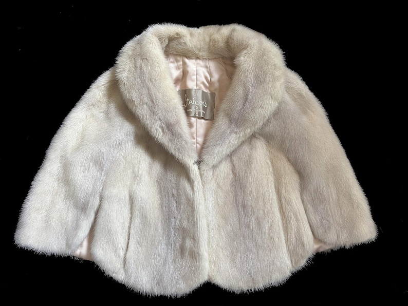 May include: A cream-coloured fur capelet featuring a wide collar and a smooth, light-coloured satin lining. A rectangular label with the text "Teigen's" is stitched into the inner neckline. The garment has a short, rounded silhouette with soft texture.