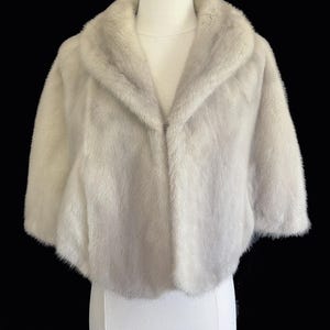 May include: A light grey, short-sleeved fur bolero jacket featuring a wide, rounded collar and a soft, dense texture. The garment is displayed on a white mannequin against a solid black background, showcasing a cropped silhouette and smooth finish.