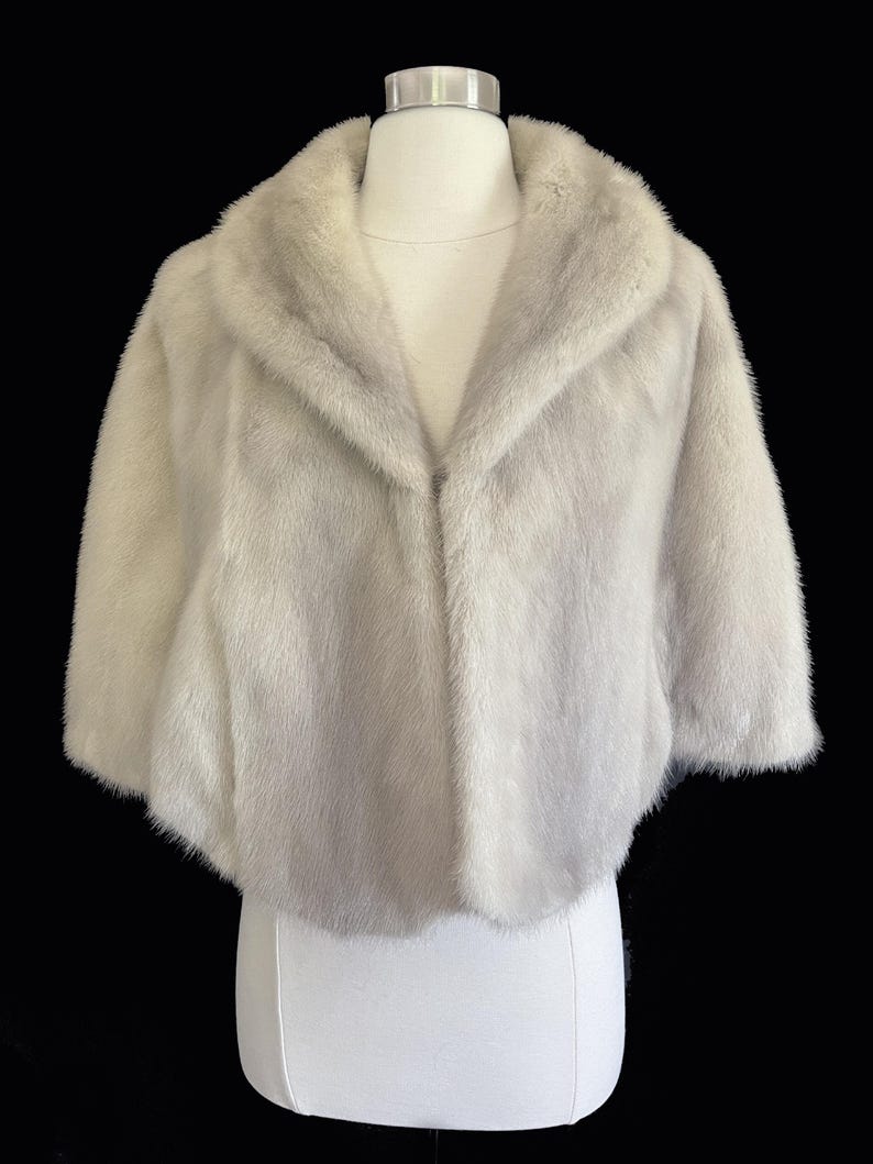 May include: A light grey, short-sleeved fur bolero jacket featuring a wide, rounded collar and a soft, dense texture. The garment is displayed on a white mannequin against a solid black background, showcasing a cropped silhouette and smooth finish.