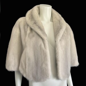 May include: A short-sleeved bolero jacket in a pale, off-white fur with a soft, dense texture. The garment features a wide, rounded shawl collar that frames the neckline and an open front design, providing a cropped silhouette that ends at the waist.