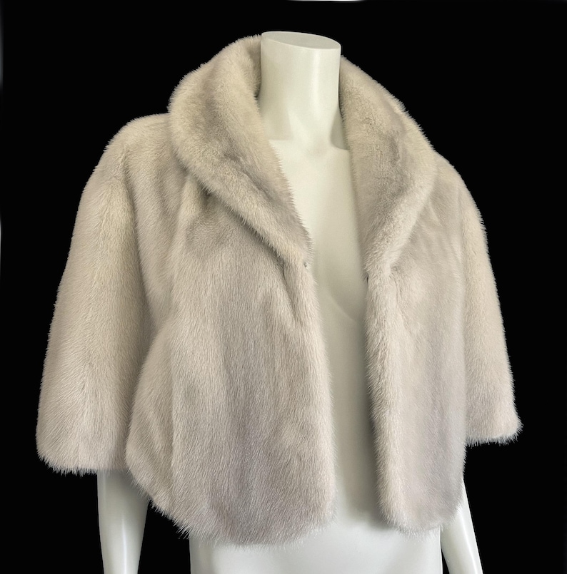 May include: A short-sleeved bolero jacket in a pale, off-white fur with a soft, dense texture. The garment features a wide, rounded shawl collar that frames the neckline and an open front design, providing a cropped silhouette that ends at the waist.