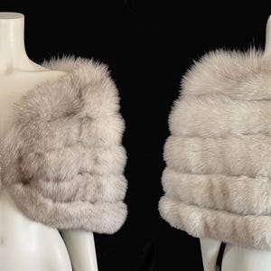 May include: A luxurious, light gray fur capelet displayed on a mannequin. The capelet features horizontal bands of soft fur, creating a layered effect. The image shows both the front and back views of the elegant garment.