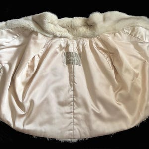 May include: A white fur bolero jacket featuring a smooth, cream-coloured satin lining. The interior displays a rectangular fabric label with the text "Jacques" stitched at the centre. The garment has short, wide sleeves and a soft, plush texture.