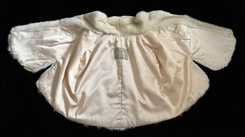 May include: A white fur bolero jacket featuring a smooth, cream-coloured satin lining. The interior displays a rectangular fabric label with the text "Jacques" stitched at the centre. The garment has short, wide sleeves and a soft, plush texture.