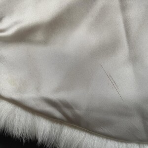 May include: Close-up of a cream-colored satin fabric with a white fur trim. The satin has a subtle sheen and a few minor imperfections. The fur is soft and fluffy, creating a contrast in textures.
