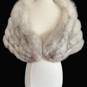 May include: A luxurious, light gray fur stole displayed on a mannequin. The fur is soft and fluffy, with a layered design. The stole is a classic accessory, perfect for formal events. The text "find your dream fur at MOXIEFURS.COM" is at the bottom.