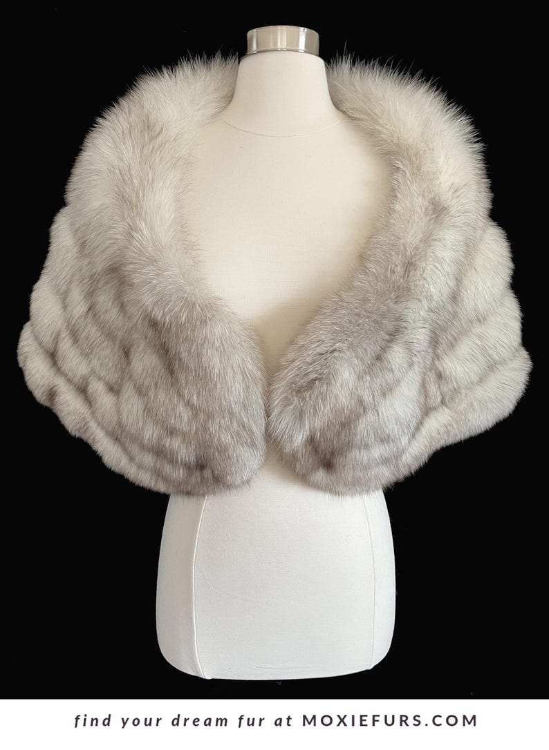 May include: A luxurious, light gray fur stole displayed on a mannequin. The fur is soft and fluffy, with a layered design. The stole is a classic accessory, perfect for formal events. The text "find your dream fur at MOXIEFURS.COM" is at the bottom.